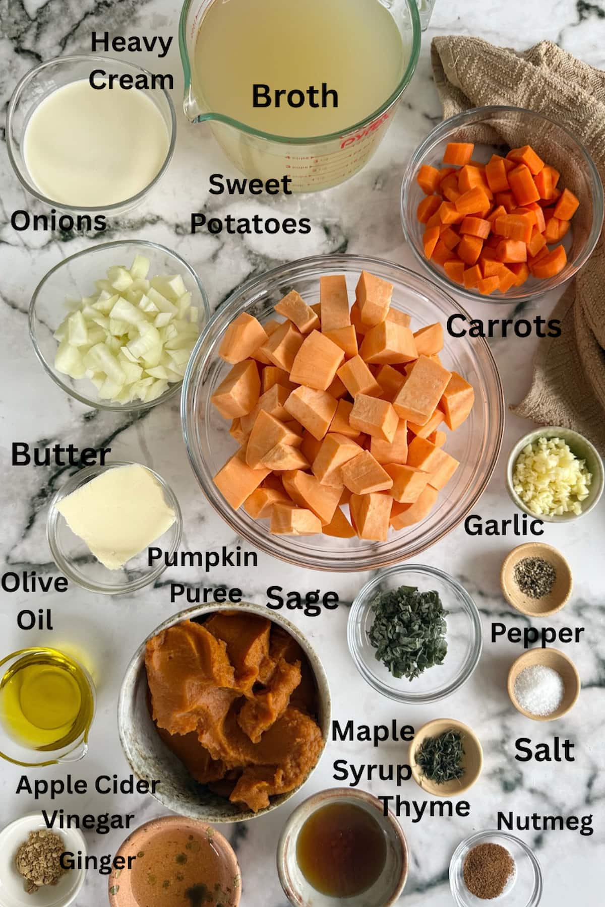 ingredients for pumpkin and sweet potato soup laid out in separate bowls - heavy cream, broth, carrots, sweet potatoes, pumpkin puree, onions, garlic, butter, olive oil, sage, thyme, pepper, salt, nutmeg, ginger, maple syrup, and apple cider vinegar.