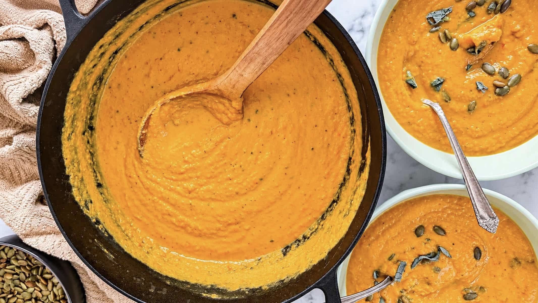 Sweet potato pumpkin soup in a cast iron skillet with two garnished bowls, pumpkin seeds, and fresh sage.