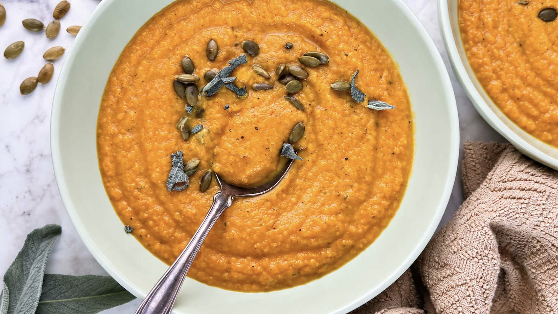 Sweet potato pumpkin soup topped with pumpkin seeds and sage, served with a spoon.