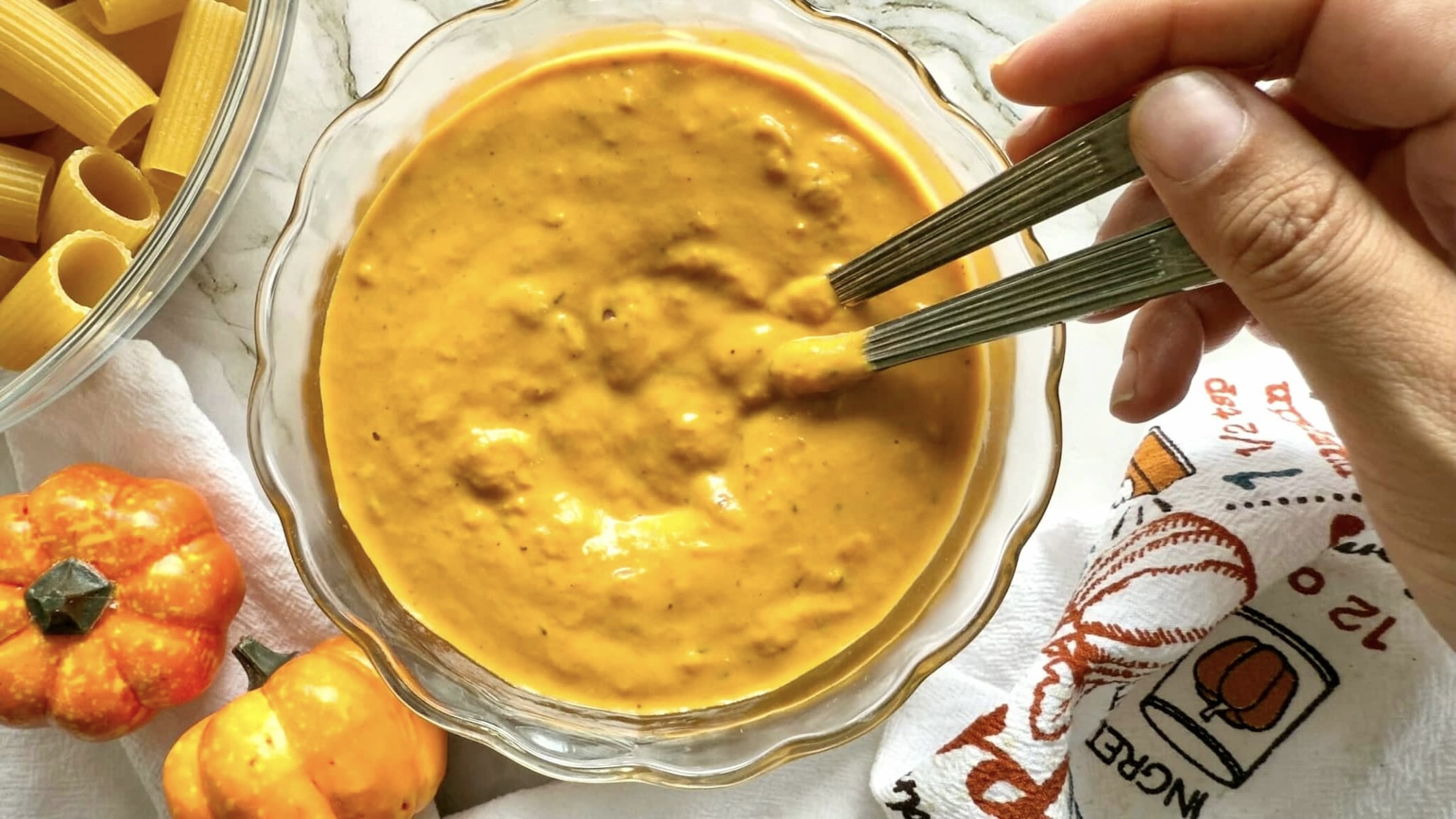 hand holding two spoons in a bowl of pumpkin sauce, bowl of dry rigatoni pasta in the background.