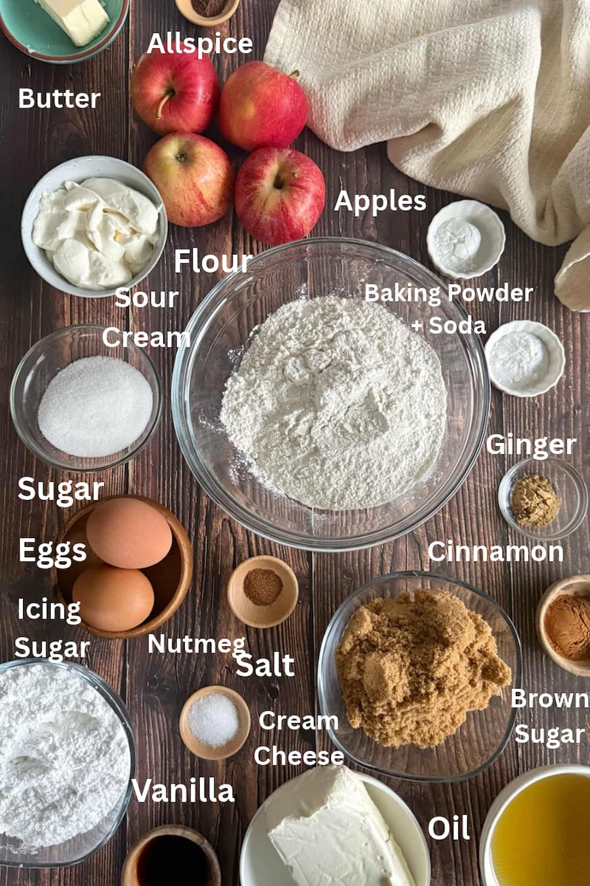 ingredients for apple spice cake laid out in separate bowls - apples, butter, allspice, sour cream, baking soda, baking powder, flour, sugar, eggs, brown sugar, icing sugar, nutmeg, ginger, vanilla, cream cheese, oil, and cinnamon.