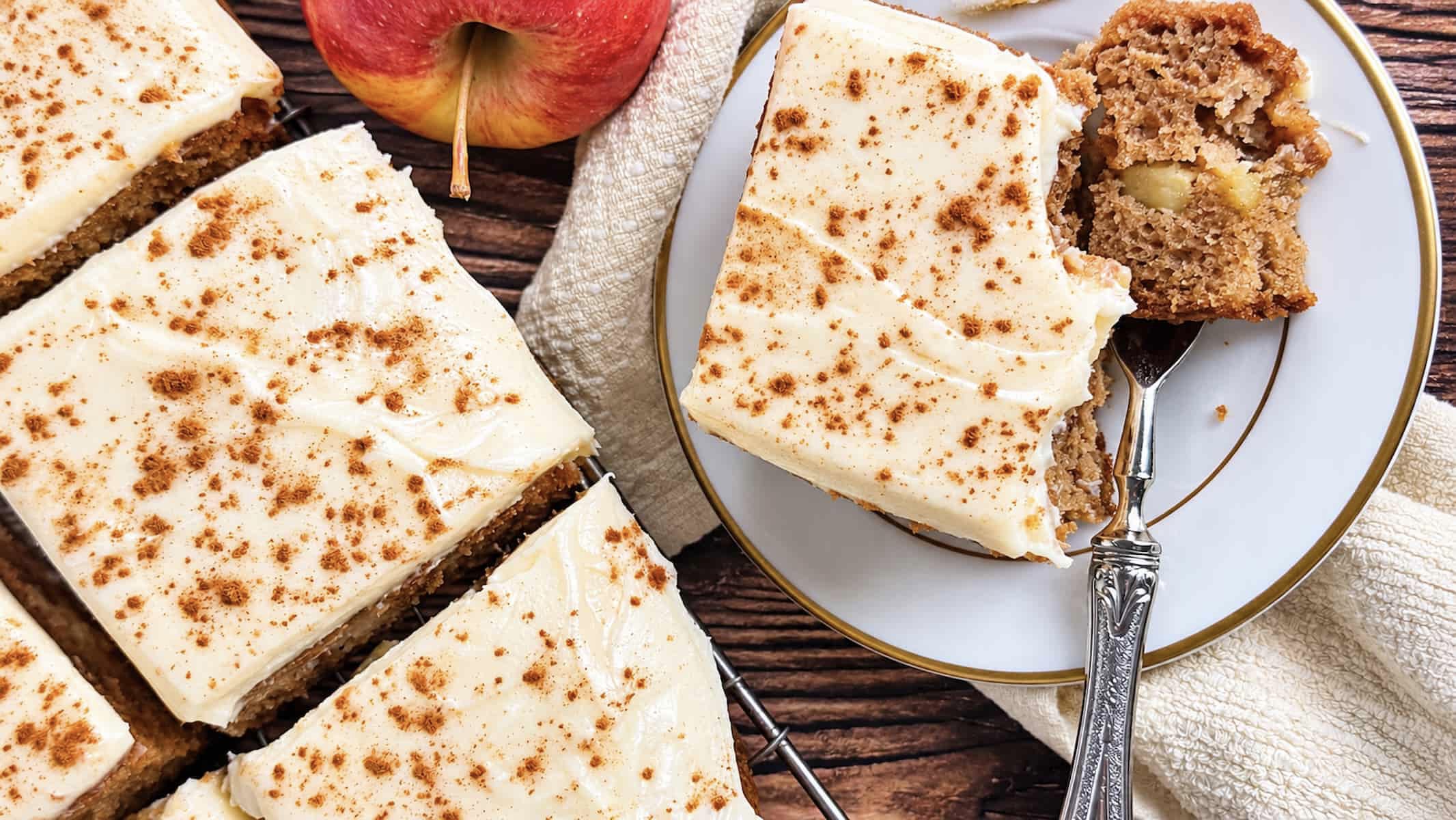 Apple spice cake squares topped with cinnamon-dusted cream cheese frosting, with a partially eaten slice on a plate beside a fresh apple.
