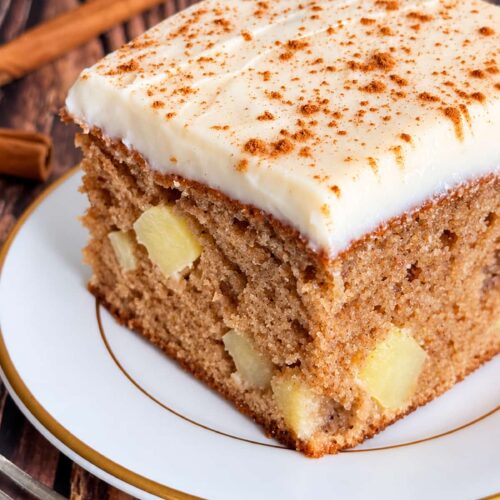 Moist apple spice cake with chunks of fresh apple, creamy cream cheese frosting, and a sprinkle of cinnamon on a white plate.