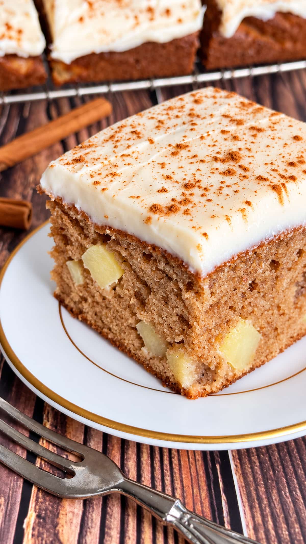 Moist apple spice cake with chunks of fresh apple, creamy cream cheese frosting, and a sprinkle of cinnamon on a white plate.