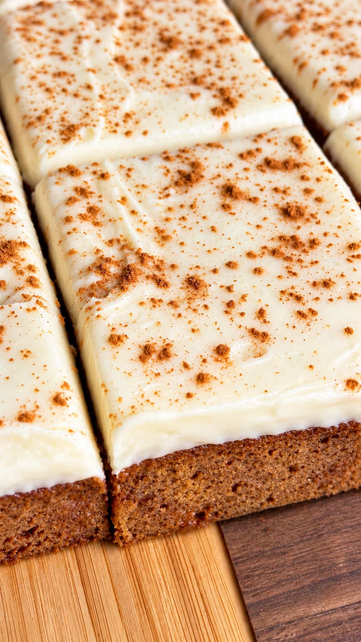 Freshly sliced apple spice cake with creamy cream cheese frosting and a sprinkle of cinnamon on a wooden cutting board.