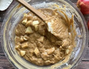 brown batter with apple chunks in a large glass bowl with a rubber spatula.