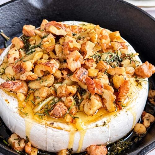 Honey baked Camembert topped with toasted walnuts, rosemary, and herbs in a cast-iron skillet.