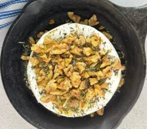 chopped walnuts and rosemary on top of a raw wheel of camembert cheese in a small cast iron skillet.