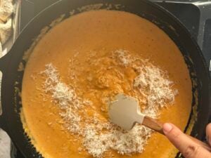 hand with a spatula stirring grated parmesan cheese into a creamy pumpkin sauce.