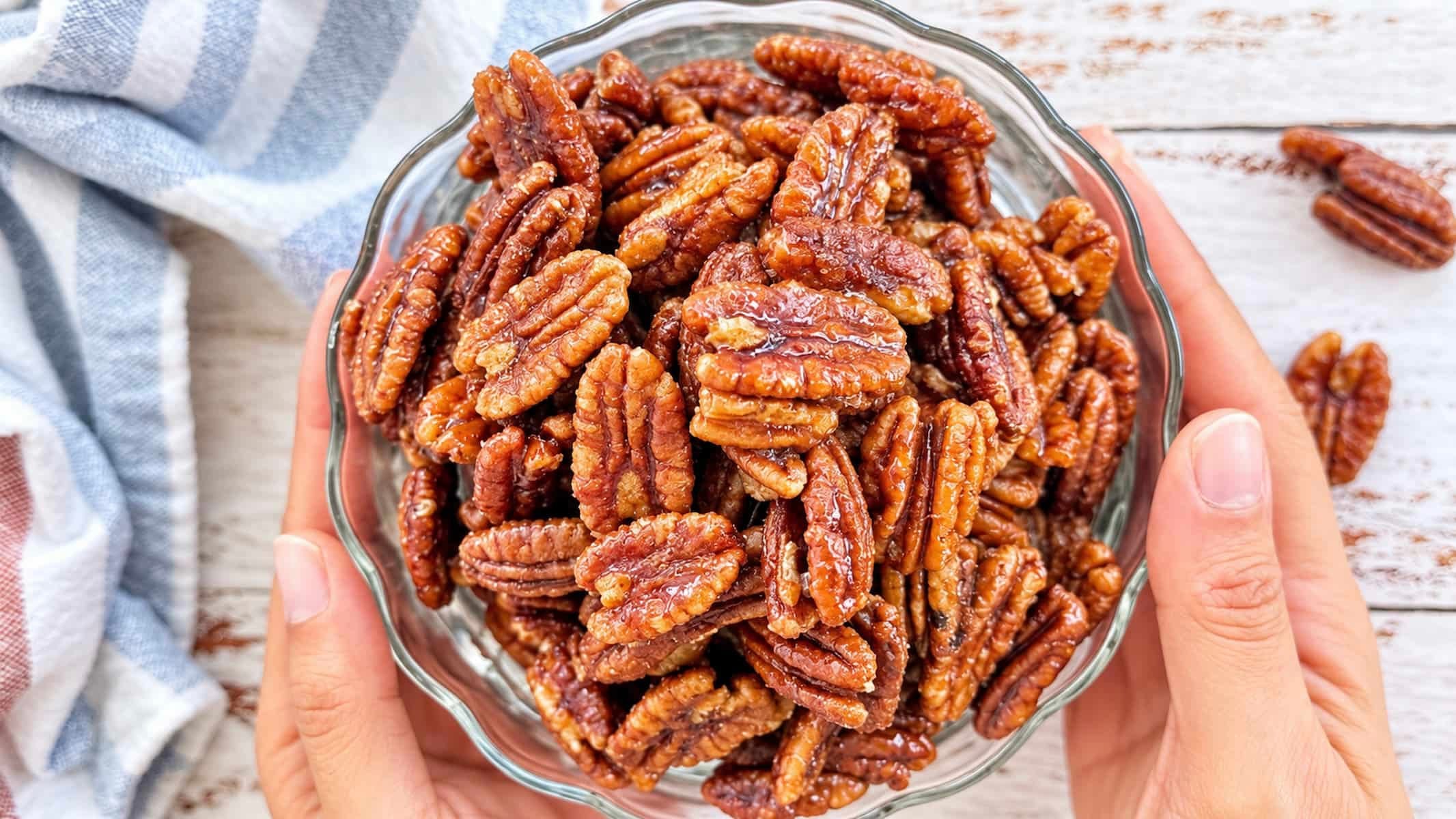 Homemade maple glazed pecans in a glass bowl, held in two hands over a rustic white wooden table.
