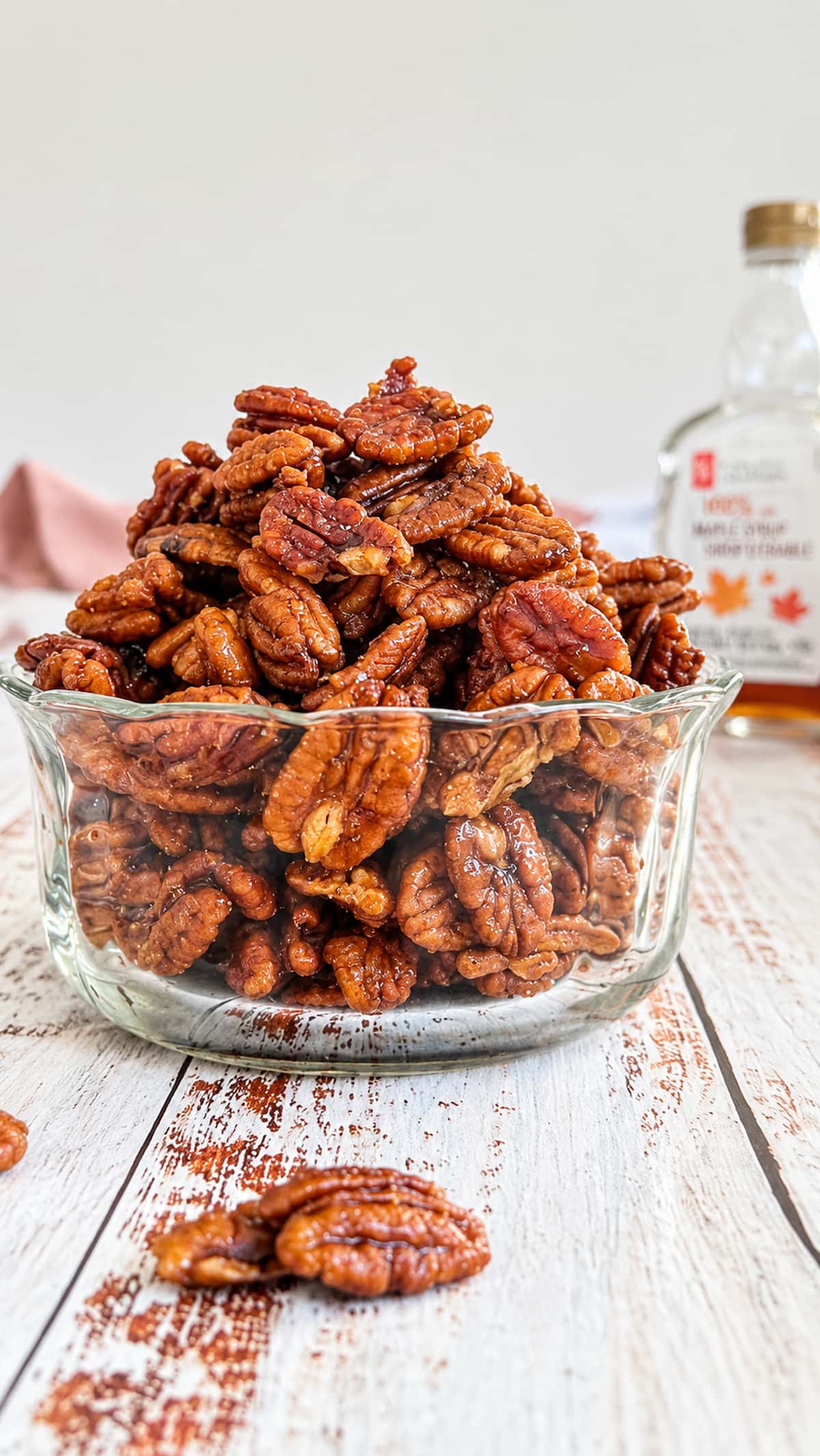 Homemade maple glazed pecans piled high in a clear glass bowl on a rustic white wooden table, with maple syrup in the background.