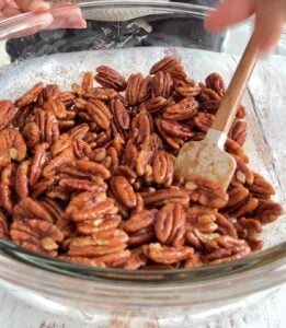 bowl of pecans tossed with maple syrup, rubber spatula in the bowl.