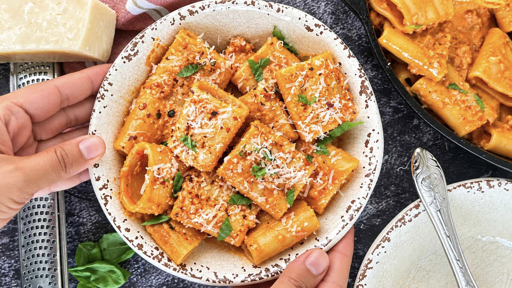 Hands holding a bowl of creamy nduja pasta with rigatoni, grated Parmesan, fresh basil, and red pepper flakes.