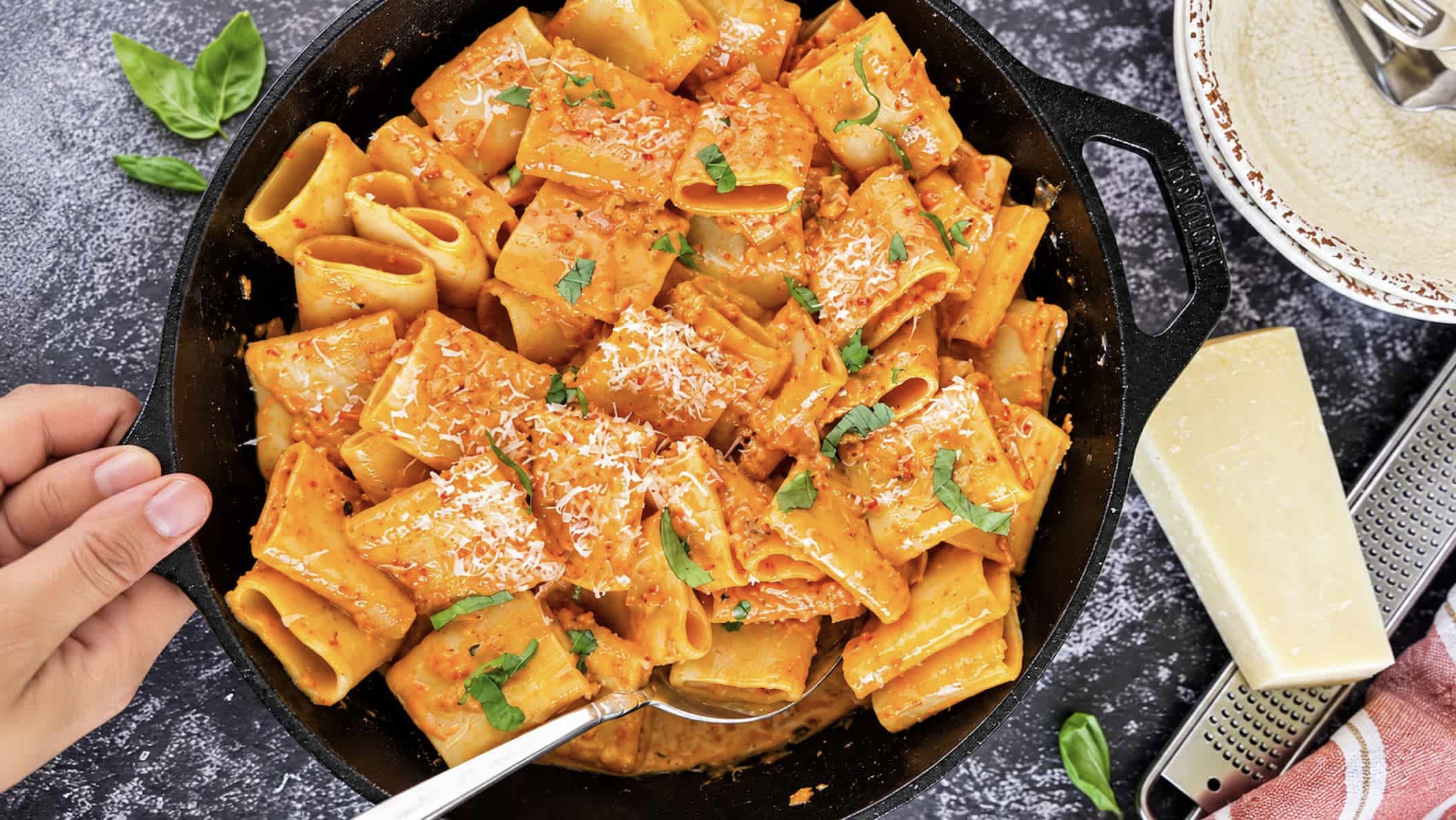 Creamy nduja rigatoni pasta with Parmesan and fresh basil in a cast iron skillet. Hands holding the handle of the skillet, block of cheese next to a microplane grater in the background.