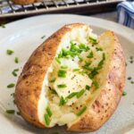 No foil baked potato with butter, fresh chives, salt, and black pepper on a plate in front of a wire rack lined baking sheet with more.