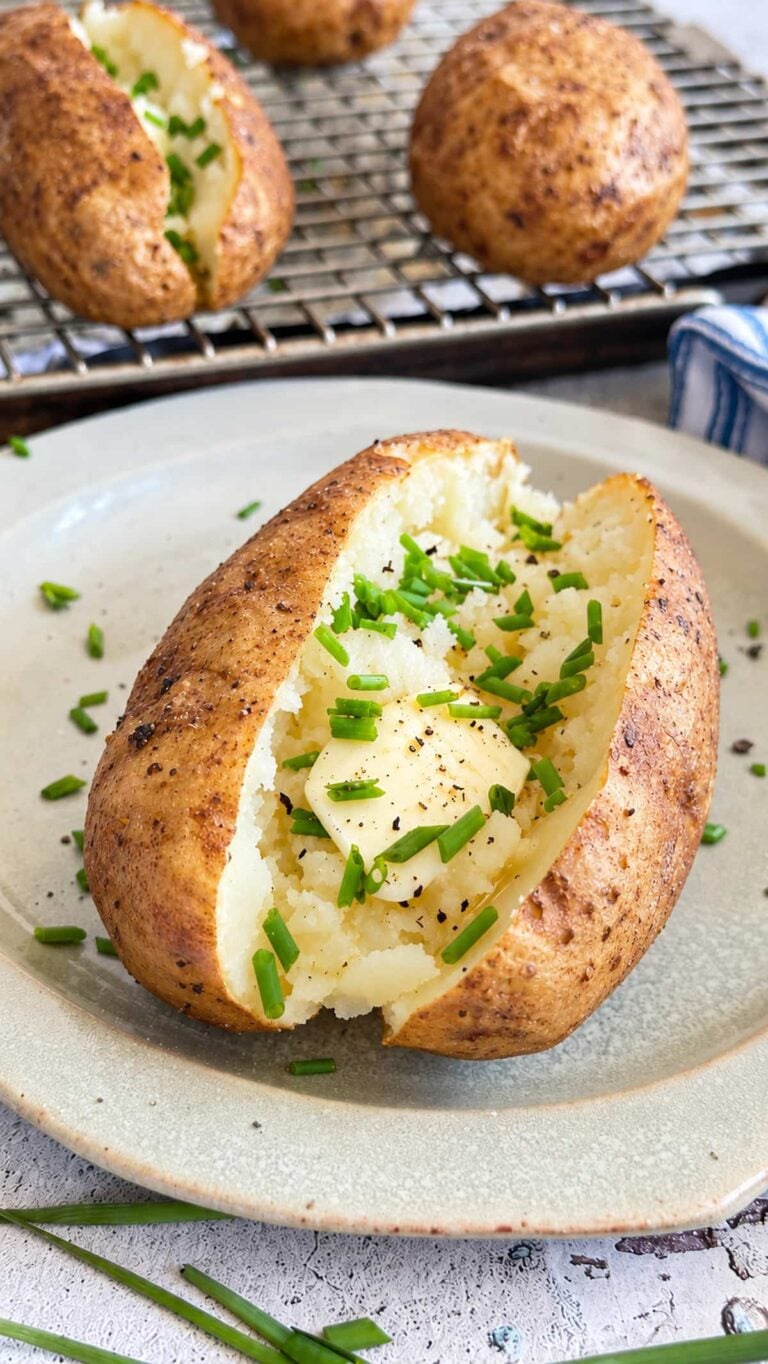 No foil baked potato with butter, fresh chives, salt, and black pepper on a plate in front of a wire rack lined baking sheet with more.