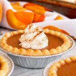 Mini pumpkin pie tarts with golden flaky crusts and creamy pumpkin filling, some topped with cinnamon-dusted whipped cream, on a baking sheet with fall pumpkins in the background.