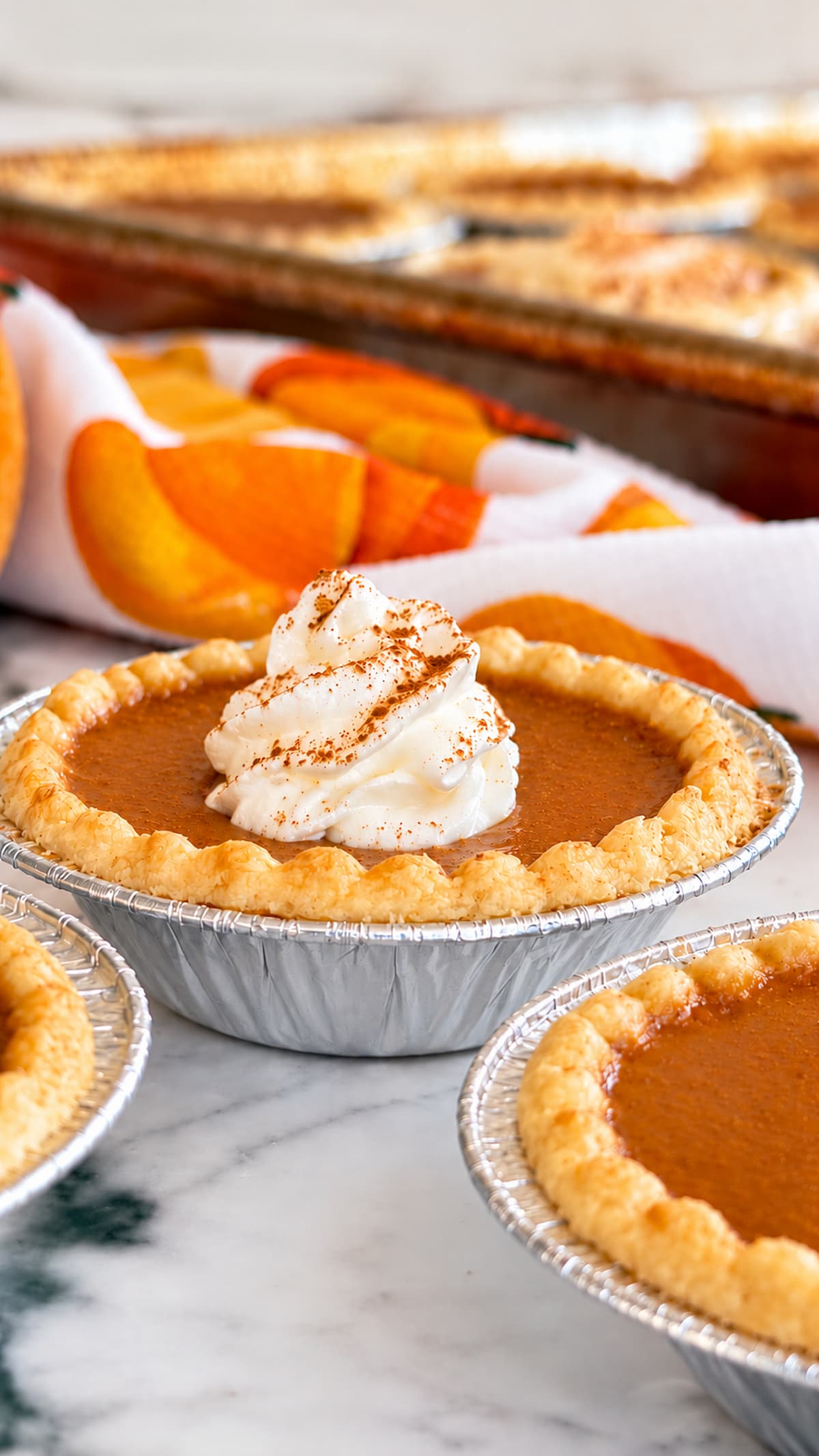Mini pumpkin pie tarts with golden flaky crusts and creamy pumpkin filling, some topped with cinnamon-dusted whipped cream, on a baking sheet with fall pumpkins in the background.