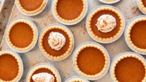 Mini pumpkin pie tarts with smooth pumpkin filling and golden flaky crusts, topped with cinnamon-dusted whipped cream on a baking sheet.