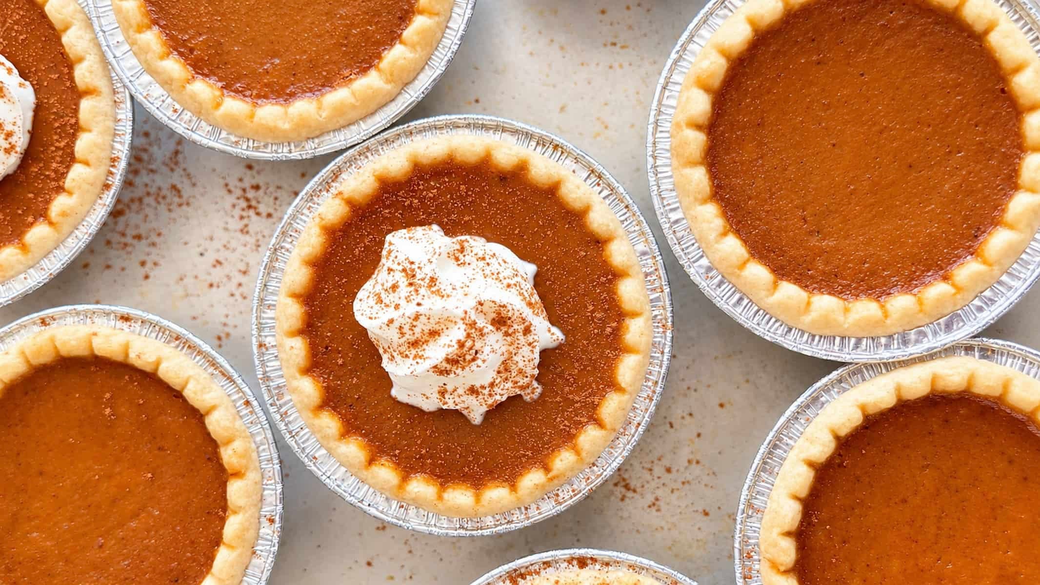 Mini pumpkin pie tarts with smooth pumpkin filling and golden flaky crusts, some topped with cinnamon-dusted whipped cream, arranged on a baking sheet.