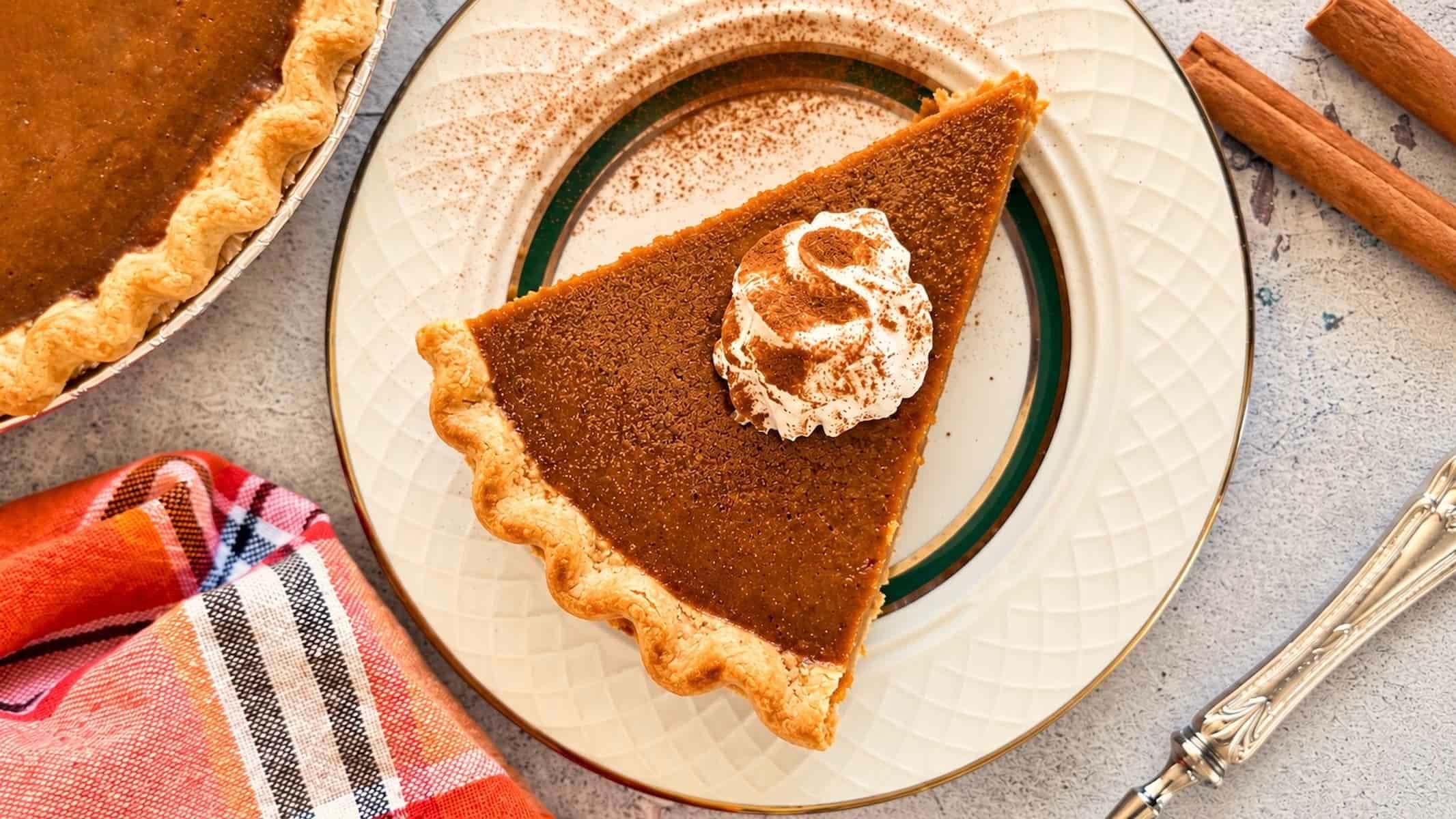 Homemade pumpkin pie slice with a golden crimped crust, topped with cinnamon-dusted whipped cream on a decorative plate.